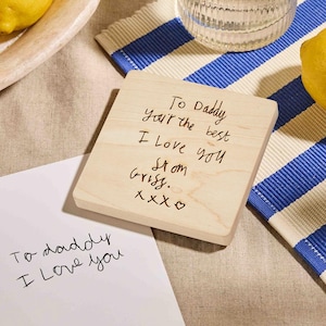 May include: A square wooden coaster with handwritten text that reads "To Daddy, you're the best, I Love you, From Grissy. xoxo". A white card with "To daddy I Love you" is also visible. The coaster is on a blue and white striped placemat.