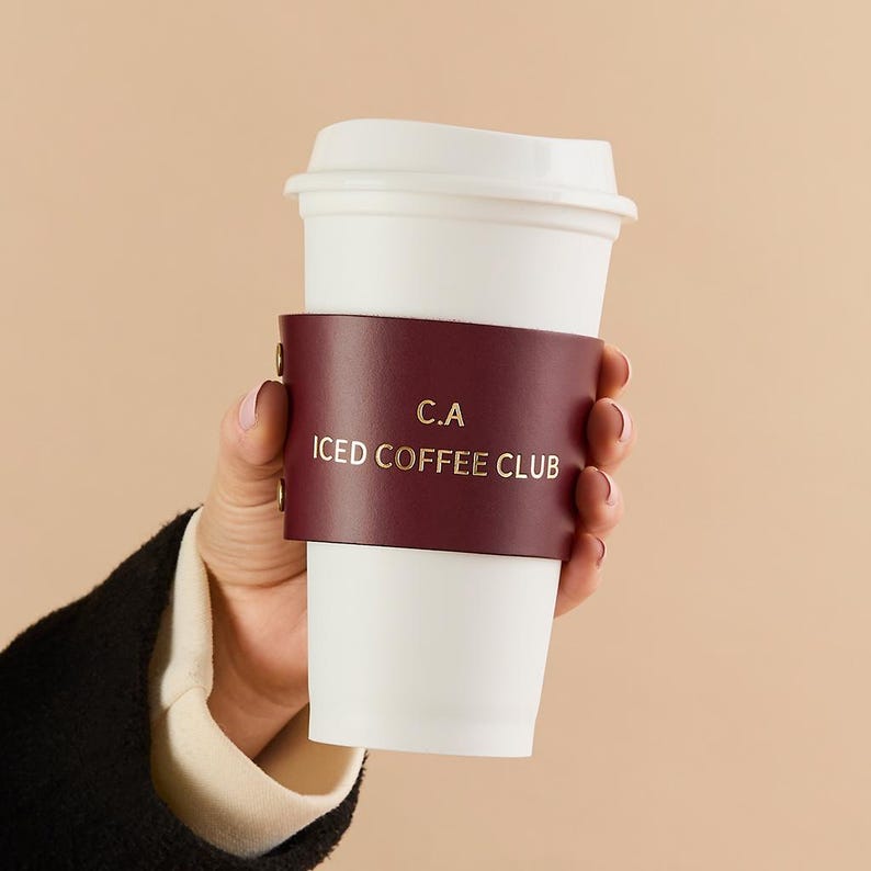 May include: A white paper coffee cup with a burgundy leather sleeve. The sleeve has gold lettering that reads "C.A ICED COFFEE CLUB". The cup is held by a person.