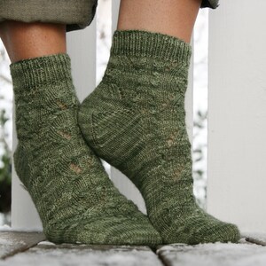 May include: A pair of green knitted socks with a lacy pattern. The socks are worn on a person's feet and are shown against a white picket fence.