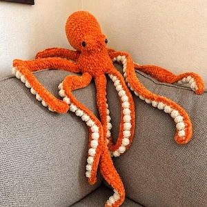 Peut inclure: Une peluche pieuvre fabriquée en fil orange vif avec une texture douce et duveteuse. La pieuvre présente une tête arrondie, deux petits yeux noirs et huit longs tentacules étendus. Chaque tentacule est bordé de petites sphères crème.