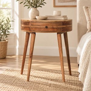 May include: A semi-circular wooden side table with four tapered legs and a single drawer featuring a small black knob. The tabletop holds a light-colored ceramic vase with greenery, a small shallow bowl, and a stack of two books with a ribbed cup.