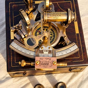 Może przedstawiać: Mosiężny sekstant z grawerowaną skalą i kompasem, oznaczony napisem J. SCOTT LONDON 1753. Instrument spoczywa na ciemnej drewnianej skrzynce ze złotymi inkrustacjami, w towarzystwie dwóch małych okrągłych mosiężnych soczewek na pierwszym planie.