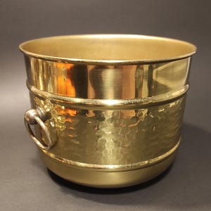 Op de afbeelding: Een emmer van gepolijst messing met een gehamerde textuur op de onderste helft en een gladde bovenrand. De houder is voorzien van twee ronde metalen ringhandvatten aan de zijkanten, wat een functionele grip biedt voor dit metalen vat.