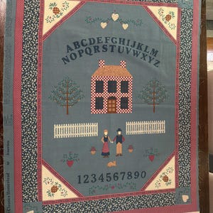 May include: A rectangular fabric panel featuring a blue-gray center with a house, a couple, a cat, and a white fence. The alphabet and numbers 0-9 are printed in dark blue. The border has a floral pattern with pineapples and hearts in the corners.