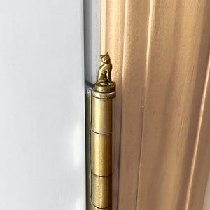 May include: A gold-toned metal door hinge featuring a small, sculpted cat figure perched on the top finial. The hinge has a cylindrical barrel design and is attached to a light-colored wooden door frame, showcasing a metallic finish throughout.