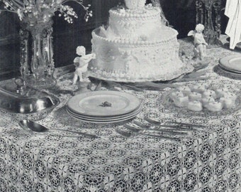 May include: A white three-tiered wedding cake decorated with white frosting and pearl-like accents sits on a white crocheted tablecloth. The cake is surrounded by white flowers and two cherub figurines.