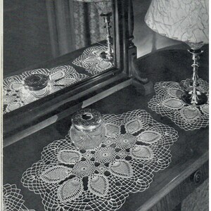 May include: Three crocheted doilies, one oval and two round, are displayed on a dark wooden surface. The doilies are made with a delicate white thread and feature a repeating pattern of leaves.