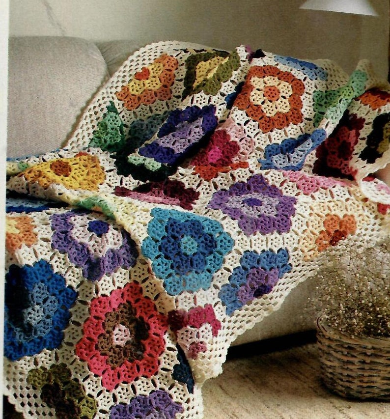 May include: A colorful crocheted throw blanket with a floral pattern. The blanket is made of white yarn with multi-colored flowers. The blanket is draped over a couch.