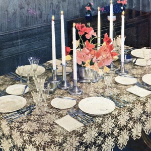 May include: A white crocheted tablecloth with a delicate floral pattern, set for a formal dinner with white plates, silverware, and glasses. A bouquet of pink and red flowers with lit candles in silver candle holders are the centerpiece.