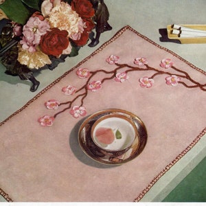 May include: A pink crocheted table runner with a brown crocheted border and a brown crocheted branch with pink flowers. The runner is on a light blue surface with a white saucer and a teacup on top of it.