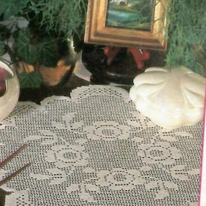 May include: A white crocheted doily with a delicate floral pattern. The doily is laid out on a dark surface with a white ceramic object in the background.