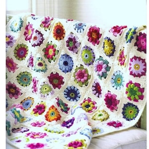 May include: A handmade crochet blanket featuring a floral pattern. The blanket is made of white yarn and adorned with colorful flower motifs in shades of pink, blue, yellow, and green. The blanket is draped over a white cushion.