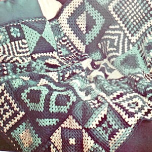 Może przedstawiać: Koc wykonany na szydełku z geometrycznym wzorem w odcieniach zieleni, bieli i czerni.