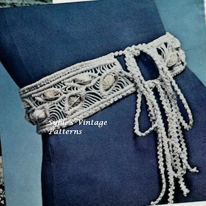 Op de afbeelding: Een witte gehaakte riem met schelpen die in het ontwerp zijn geweven. De riem is aan de voorkant in een strik geknoopt. De riem bevindt zich op een blauwe achtergrond. "Syble's Vintage Patterns" is op de afbeelding gedrukt.