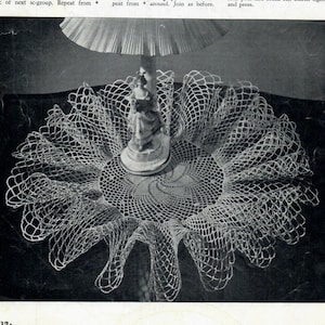 May include: A white crocheted doily with a scalloped edge, featuring a delicate spiral design in the center. The doily is displayed on a table with a decorative figurine.