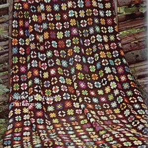 May include: A brown crochet blanket with a colorful floral pattern. The blanket is made of squares with a flower design in the center. The flowers are in various colors, including red, yellow, blue, green, and white. The blanket is draped over a wooden fence.