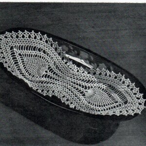 May include: A white crocheted oval doily with a delicate, lacy design. The doily is placed on a dark surface, creating a striking contrast.