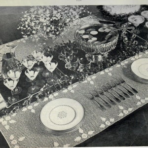 May include: A white crocheted table runner with a delicate pattern. The runner is laid on a table with a glass pitcher, glasses, plates, and cutlery. There is a bowl of fruit in the background.