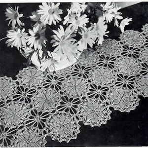 May include: A black and white photograph of a handmade, lacy table runner with a floral design. The runner is placed on a dark surface, and a vase of white daisies sits in the background.