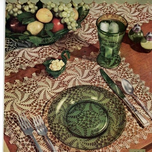 May include: A table setting with a green glass plate and a white crocheted doily. The table is set with a green glass plate, a silver knife and spoon, and two silver forks. There is a green glass cup with ice cubes and two small glass salt shakers on the table.