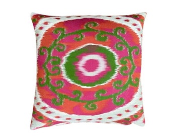 May include: A square pillow with a white background and a large, colorful, circular design. The design features a pink and white center surrounded by a green ring, all within a red and pink ring. The outer ring is decorated with green floral patterns.