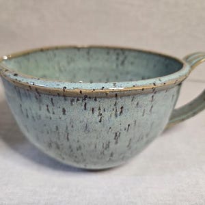 May include: A ceramic mixing bowl with a light blue-green glaze and dark brown speckles throughout. The bowl features a small pouring spout on the left side and a curved handle on the right side, with a smooth, rounded rim and a deep basin.