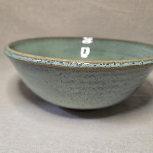 May include: A round ceramic bowl with a light mint green glaze and a speckled texture. The rim of the bowl features a darker brown, earthy tone with visible flecks of blue and grey, creating a contrast against the smooth interior surface.