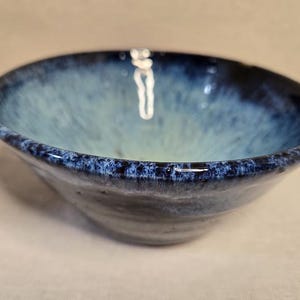 May include: A ceramic bowl featuring a deep blue rim that transitions into a lighter, pale blue center. The glaze has a mottled, speckled appearance with a smooth, rounded edge and a slightly tapered base, creating a simple, functional vessel.