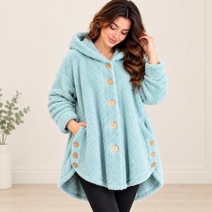 Könnte beinhalten: Eine hellblaue Kapuzenjacke aus strukturiertem Fleece-Material. Die Jacke verfügt über eine Knopfleiste vorne mit fünf großen, runden Holzknöpfen sowie drei zusätzlichen Holzknöpfen auf jeder Seite in der Nähe der Taschen am Saum.