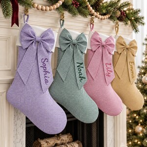 May include: Four Christmas stockings in lavender, sage green, pink, and tan gingham fabric, each featuring a large matching bow and a white cuff. The stockings are personalized with the embroidered names Sophia, Noah, Lily, and Emma.
