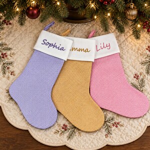 May include: Three Christmas stockings featuring a small gingham check pattern in purple, yellow, and pink. Each stocking has a white cuff with a name embroidered in a matching color: Sophia in purple, Emma in gold, and Lily in pink.