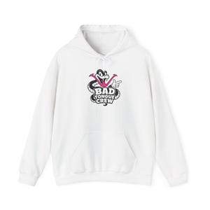 Puede incluir: Una sudadera con capucha blanca que presenta un gráfico en el pecho de una serpiente de dibujos animados en blanco y negro con una lengua rosa brillante. La serpiente señala a la derecha y el texto BAD TONGUE CREW está impreso en letras negras.