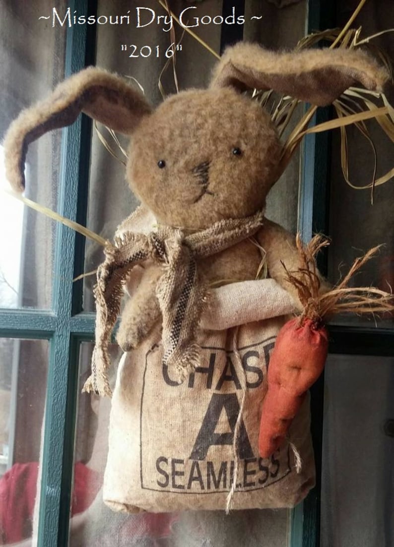 May include: A handcrafted brown bunny doll with long ears, wearing a burlap scarf and holding a fabric carrot. The bunny is holding a burlap sack with the text "CHAS A SEAMLESS". The image also includes the text "Missouri Dry Goods" and "2016".
