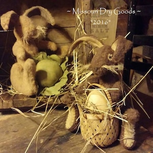 May include: Two handmade plush bunny rabbits, one brown and one tan, sitting in a nest of straw. The brown bunny is holding a basket with a white egg inside. The tan bunny is holding a green egg. The sign behind the bunnies reads "Missouri Dry Goods" and "2016".