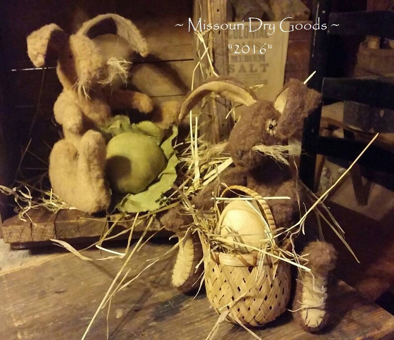 May include: Two handmade plush bunny rabbits, one brown and one tan, sitting in a nest of straw. The brown bunny is holding a basket with a white egg inside. The tan bunny is holding a green egg. The sign behind the bunnies reads "Missouri Dry Goods" and "2016".