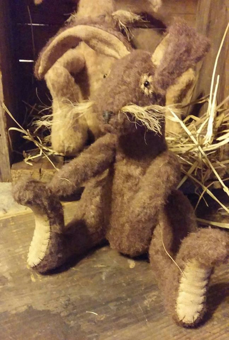 May include: A brown plush rabbit toy with long ears and a stitched nose. The rabbit is sitting on a wooden surface with straw in the background.