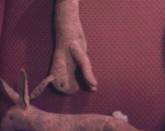 May include: Two plush rabbit toys in a light brown color are displayed on a red surface. One rabbit is upright, while the other is lying down. The toys have long ears and small tails, suggesting a whimsical theme.