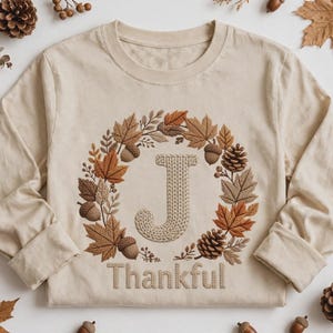 Könnte beinhalten: Ein beiges Langarmshirt mit einem mittigen, gestickten Kranz aus Herbstblättern, Tannenzapfen und Eicheln, der den Buchstaben J umgibt. Unter dem Kranz ist das Wort Thankful in einer passenden beigen, strukturierten Schrift gestickt.
