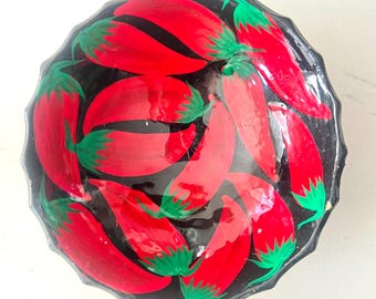 May include: A round, scalloped-edge bowl with a glossy black finish, decorated with a repeating pattern of vibrant red chili peppers featuring bright green stems arranged in a circular orientation across the interior surface.