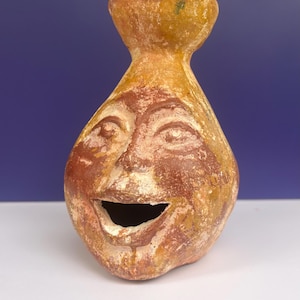 Może przedstawiać: Ceramiczne naczynie w kolorze terakoty w kształcie głowy z szerokimi, otwartymi ustami i wąską szyjką. Powierzchnia ma zwietrzałą, nakrapianą fakturę w odcieniach żółci, brązu i beżu, przedstawiającą wyrzeźbioną twarz z oczami.