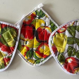 May include: Three quilted pot holders featuring a vibrant fruit pattern with red apples, yellow pears, and green leaves on a white background. Each square-shaped kitchen textile is finished with a white fabric trim and a small hanging loop.