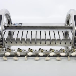 Op de afbeelding: Een roestvrijstalen zeepsnijder met een reeks verticale snijdraden, een basis met meerdere sleuven en een rij witte spanknoppen aan de voorzijde. Een klein zwart label bevat de witte tekst Soap Cutter Pro.