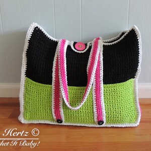 Op de afbeelding: Een zwarte, groene en witte gehaakte tote bag met roze en witte banden. De tas heeft een knoopsluiting en een gehaakte rand.