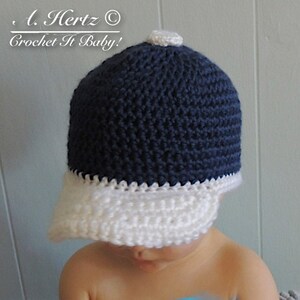 Puede incluir: Una gorra de b&eacute;isbol de crochet azul marino y blanco con un pomp&oacute;n blanco en la parte superior. La gorra tiene una visera blanca y est&aacute; hecha con un punto texturizado.