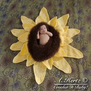 May include: A crocheted sunflower baby prop with a brown fuzzy center. A baby is lying in the center of the sunflower. The sunflower is on a green and brown floral patterned fabric.