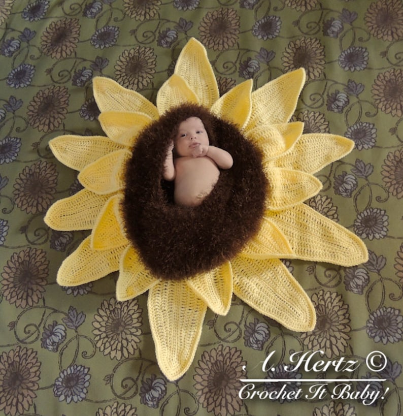 May include: A crocheted sunflower baby prop with a brown fuzzy center. A baby is lying in the center of the sunflower. The sunflower is on a green and brown floral patterned fabric.