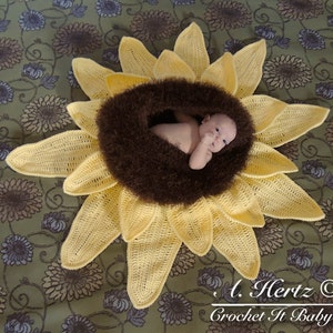 May include: A crocheted sunflower baby nest with a brown fuzzy center. A baby is lying in the nest. The text "A. Hertz &copy; Crochet It Baby!" is in the bottom right corner of the image.