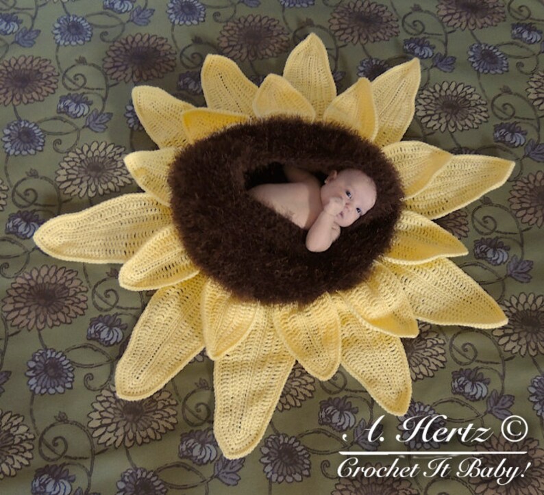 May include: A crocheted sunflower baby nest with a brown fuzzy center. A baby is lying in the nest. The text "A. Hertz &copy; Crochet It Baby!" is in the bottom right corner of the image.