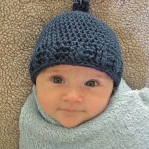 May include: A baby wearing a blue crocheted hat with a pompom on top. The hat has a textured stitch pattern. The baby is wrapped in a light blue blanket.
