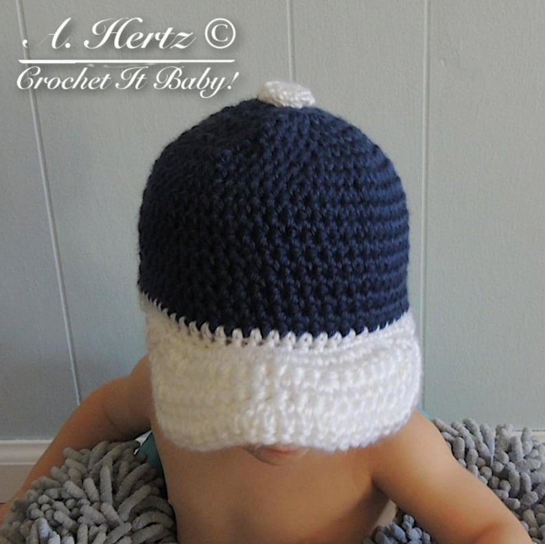 Puede incluir: Un gorro de beb&eacute; tejido a crochet azul marino y blanco con visera. El gorro tiene un pomp&oacute;n blanco en la parte superior. El texto "A. Hertz &copy; Crochet It Baby!" es visible sobre el gorro.